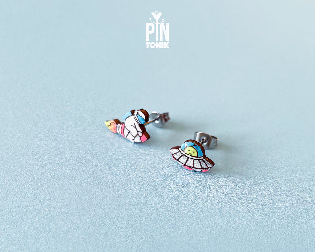 Handmade wooden mismatched stud earrings featuring an astronaut floating in space on one ear and a pastel alien UFO spaceship on the other by PinTonik.
