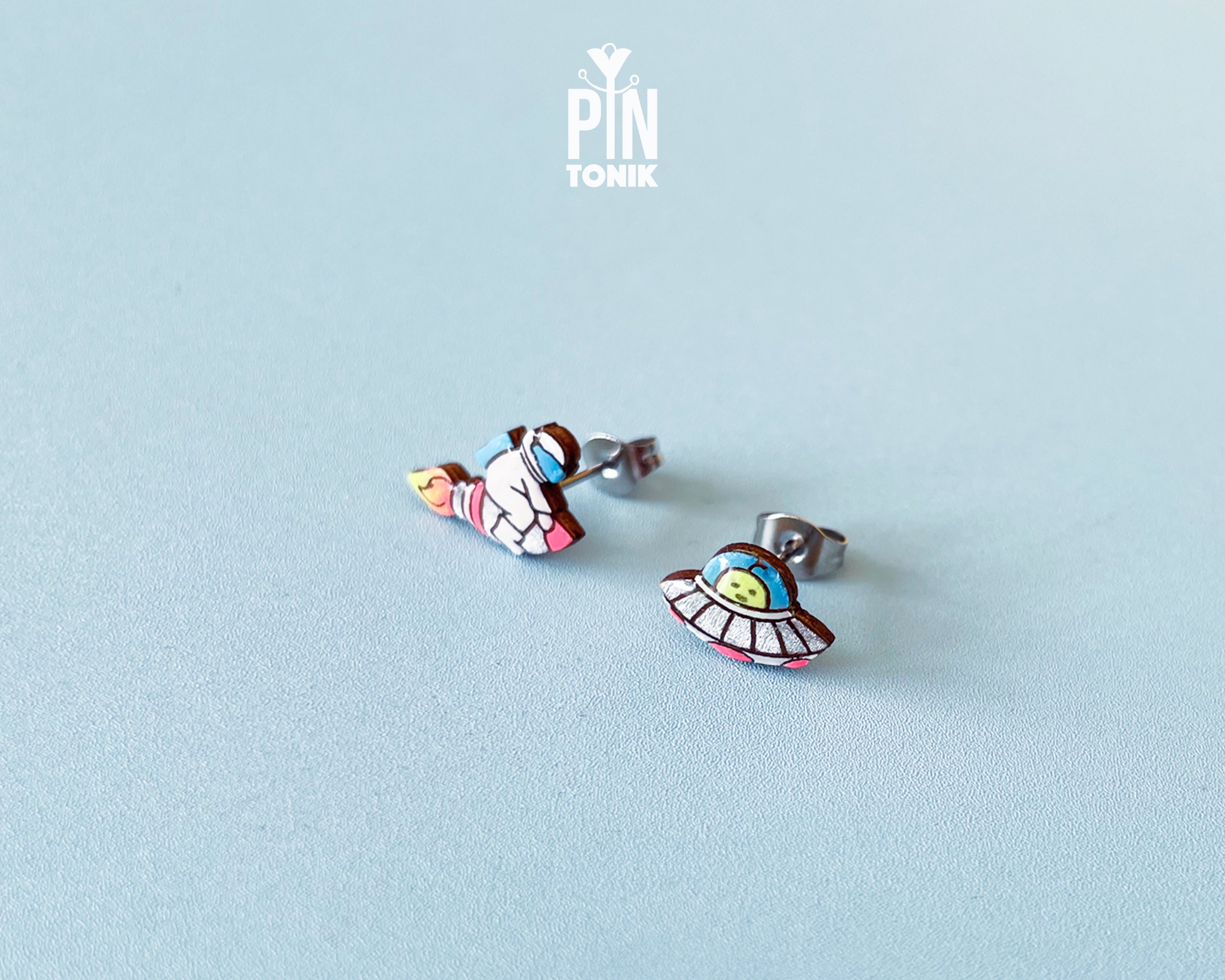 Handmade wooden mismatched stud earrings featuring an astronaut floating in space on one ear and a pastel alien UFO spaceship on the other by PinTonik.