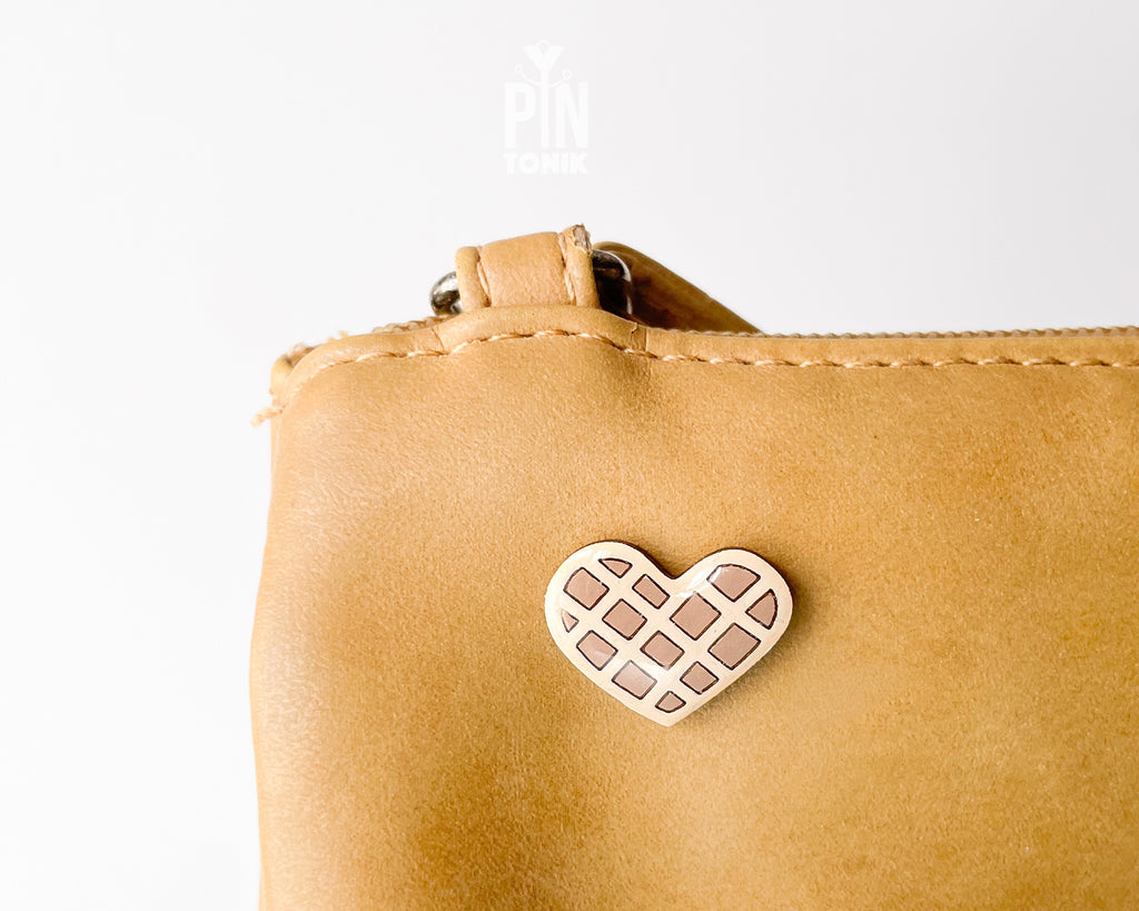 Hand-painted wooden heart-shaped waffle pin with golden-brown detailing and a glossy resin finish, shown next to a gold-toned clutch back on a light blue background.