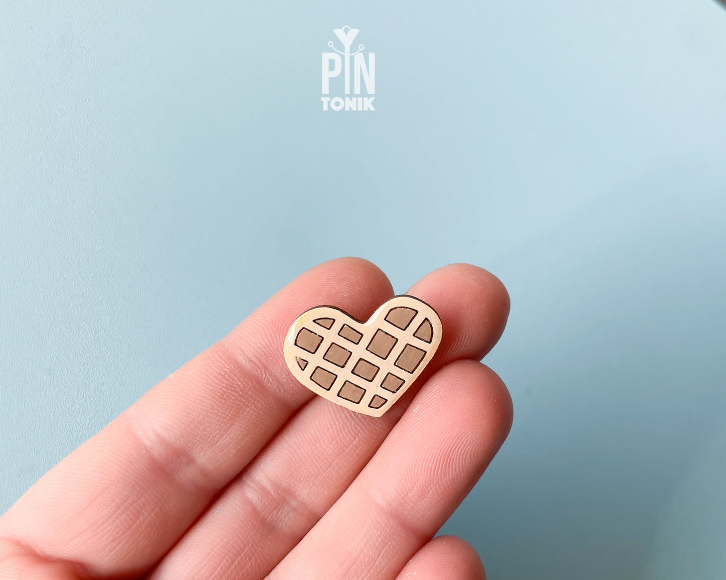 Hand-painted wooden heart-shaped waffle pin with golden-brown detailing and a glossy resin finish, shown next to a gold-toned clutch back on a light blue background.