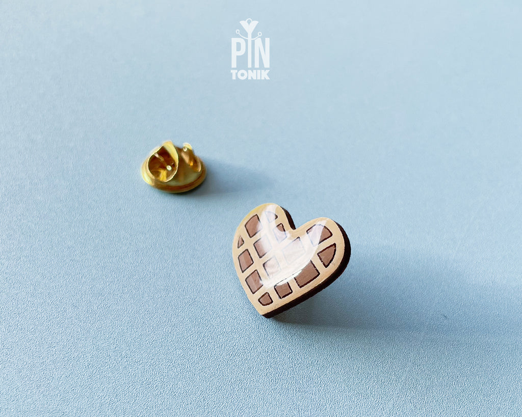 Hand-painted wooden heart-shaped waffle pin with golden-brown detailing and a glossy resin finish, shown next to a gold-toned clutch back on a light blue background.
