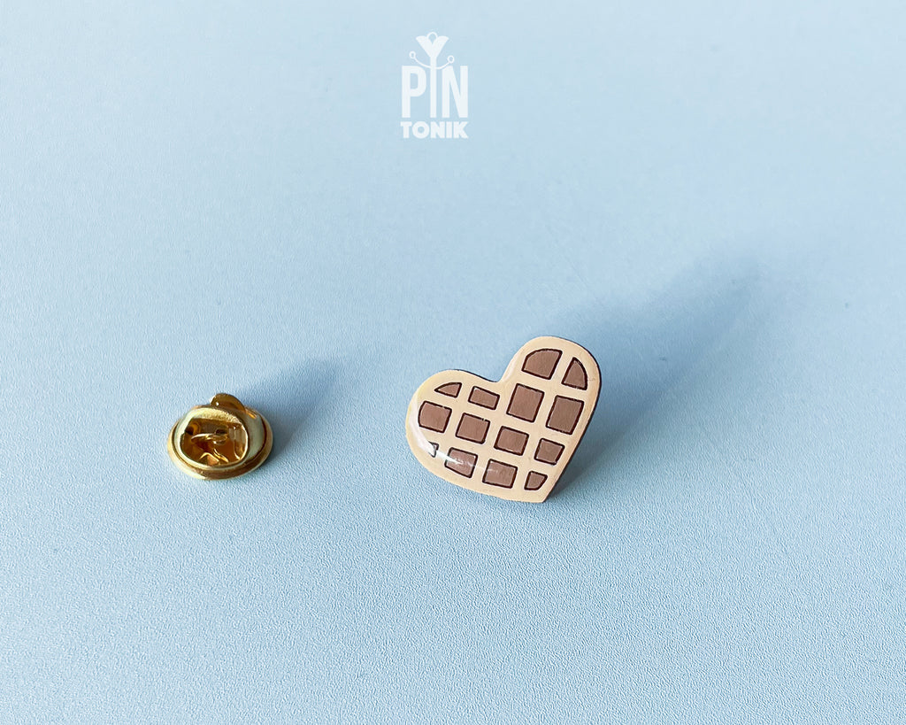 Hand-painted wooden heart-shaped waffle pin with golden-brown detailing and a glossy resin finish, shown next to a gold-toned clutch back on a light blue background.