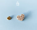 Hand-painted wooden heart-shaped waffle pin with golden-brown detailing and a glossy resin finish, shown next to a gold-toned clutch back on a light blue background.