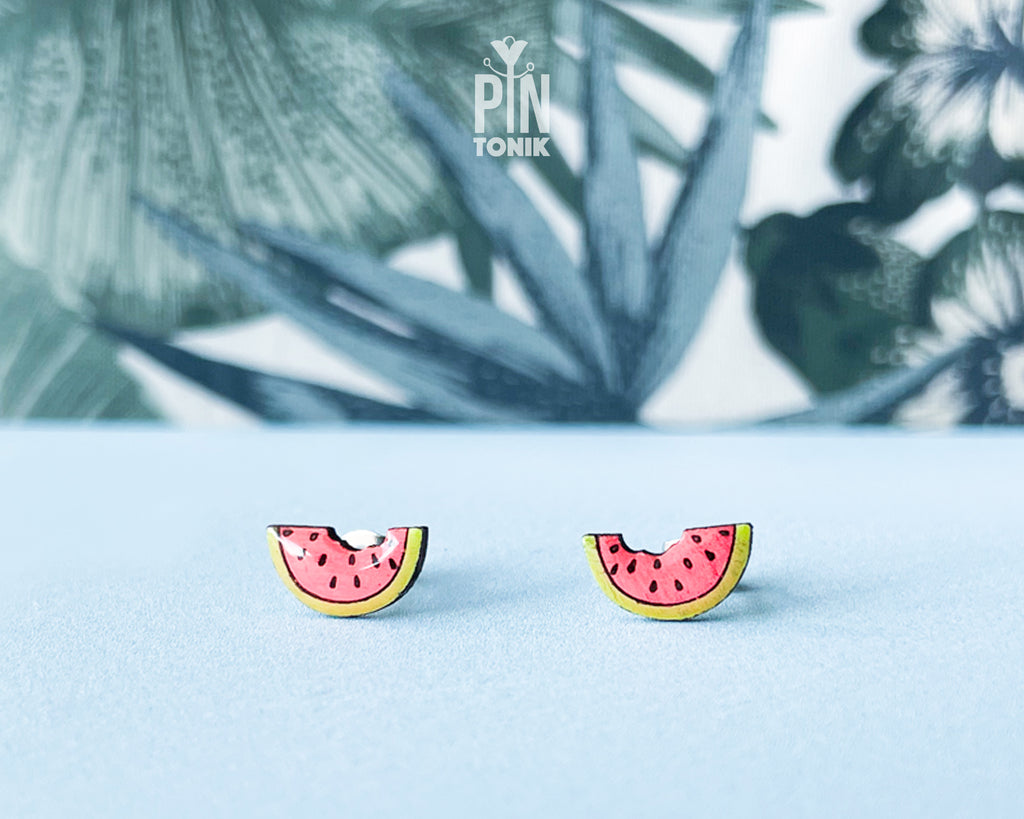 Watermelon slice wooden stud earrings with a tiny bite mark, featuring vibrant pink and green colors on a tropical leaf background.
