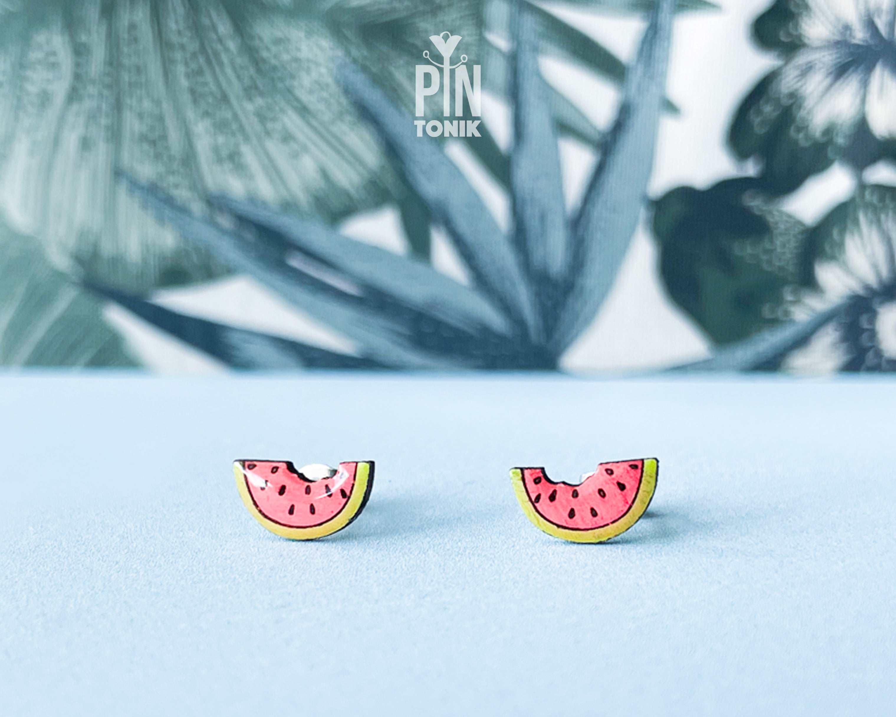 Watermelon slice wooden stud earrings with a tiny bite mark, featuring vibrant pink and green colors on a tropical leaf background.