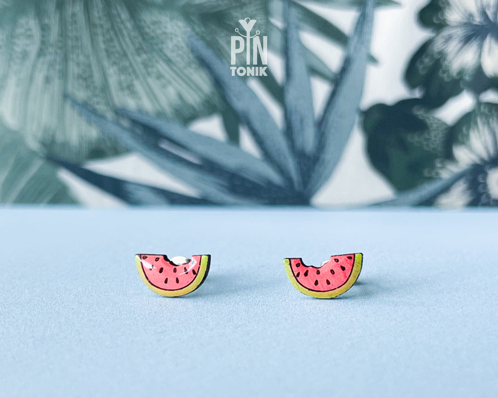 Watermelon slice wooden stud earrings with a tiny bite mark, featuring vibrant pink and green colors on a tropical leaf background.