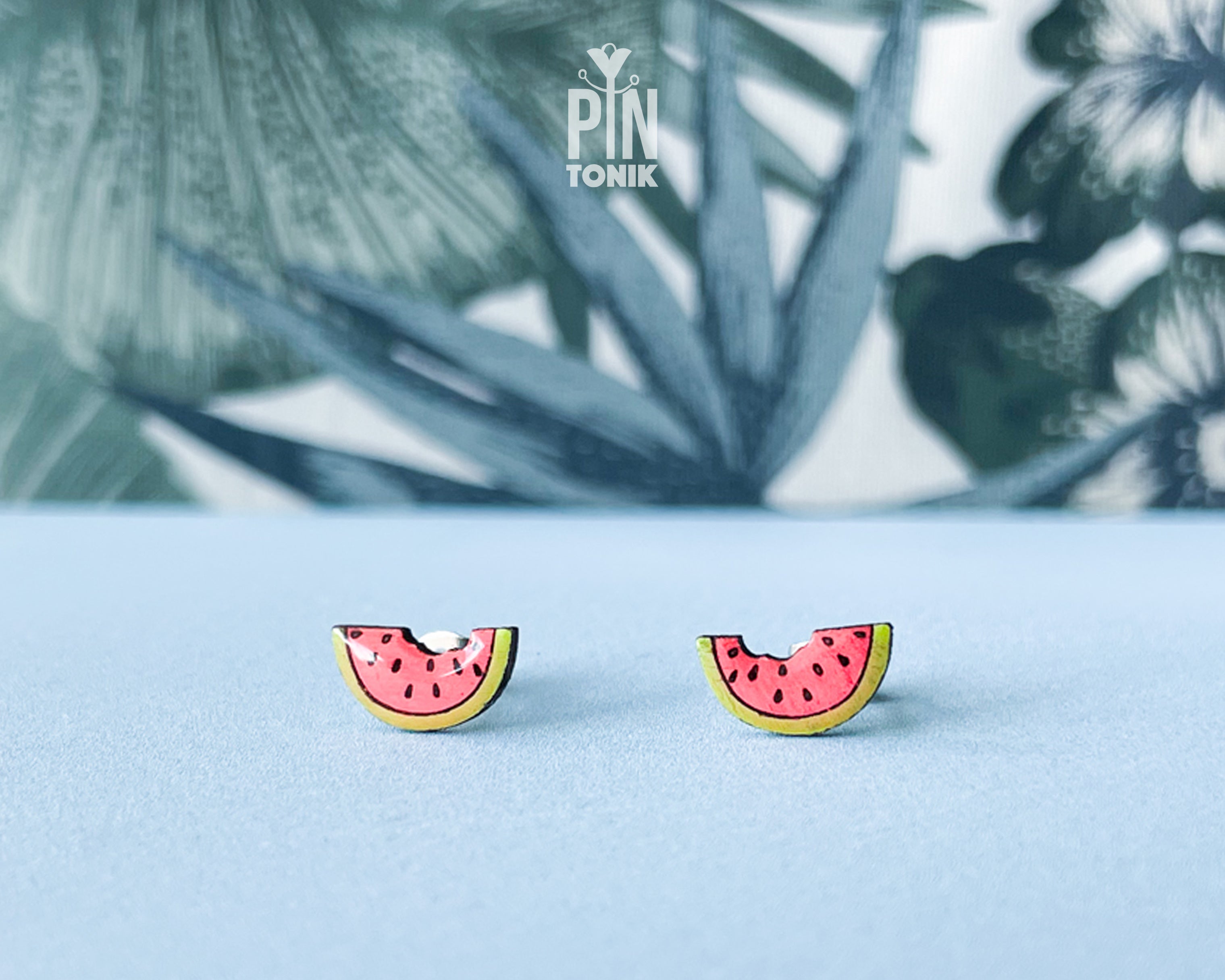Watermelon slice wooden stud earrings with a tiny bite mark, featuring vibrant pink and green colors on a tropical leaf background.