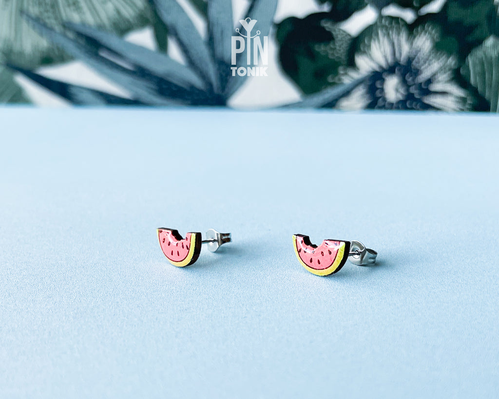 Watermelon slice wooden stud earrings with a tiny bite mark, featuring vibrant pink and green colors on a tropical leaf background.