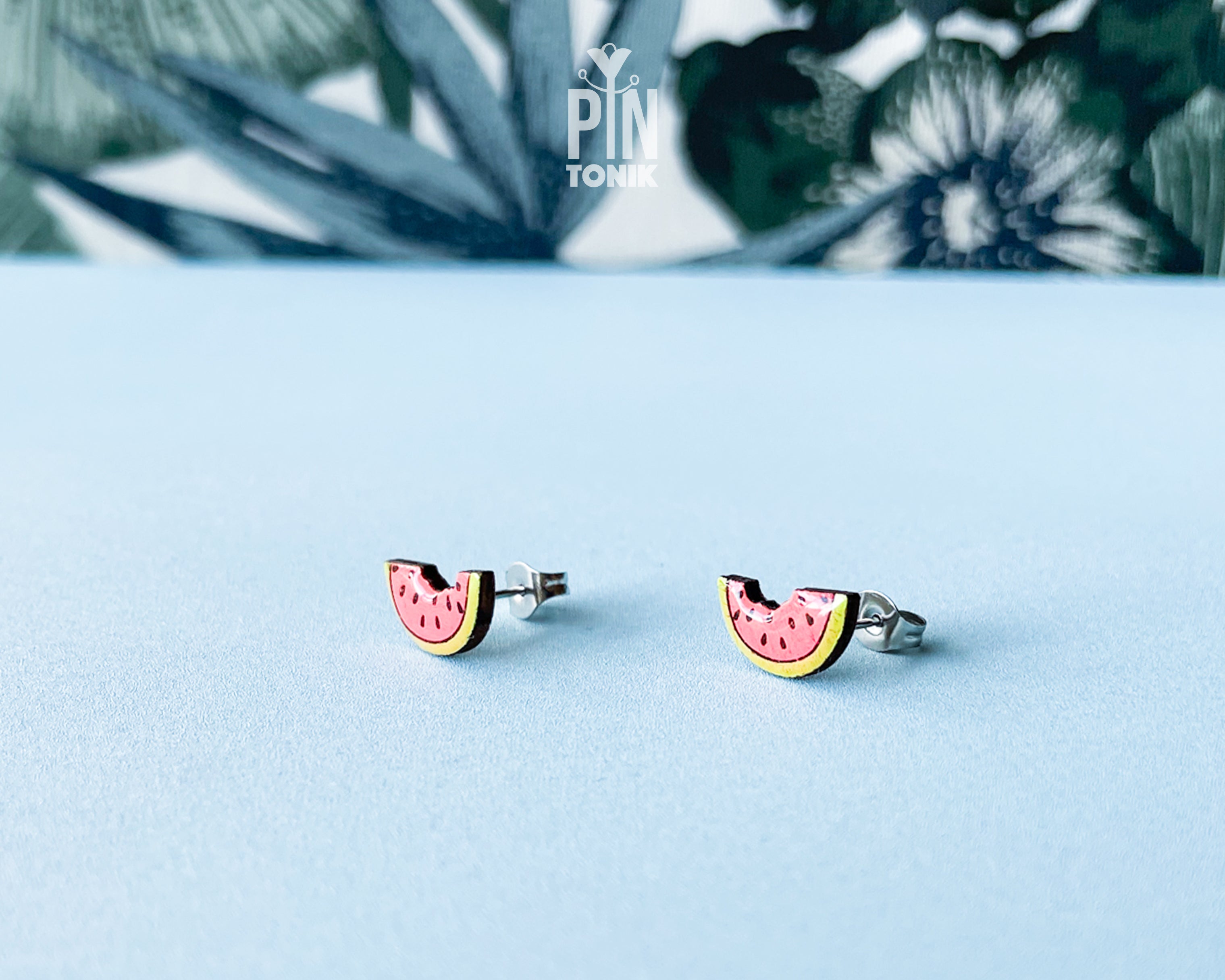 Watermelon slice wooden stud earrings with a tiny bite mark, featuring vibrant pink and green colors on a tropical leaf background.