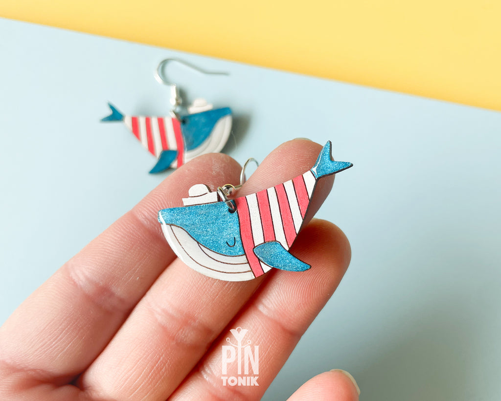 Hand-painted wooden blue whale dangle earrings wearing red and white striped sweaters and tiny sailor hats on sterling silver hooks