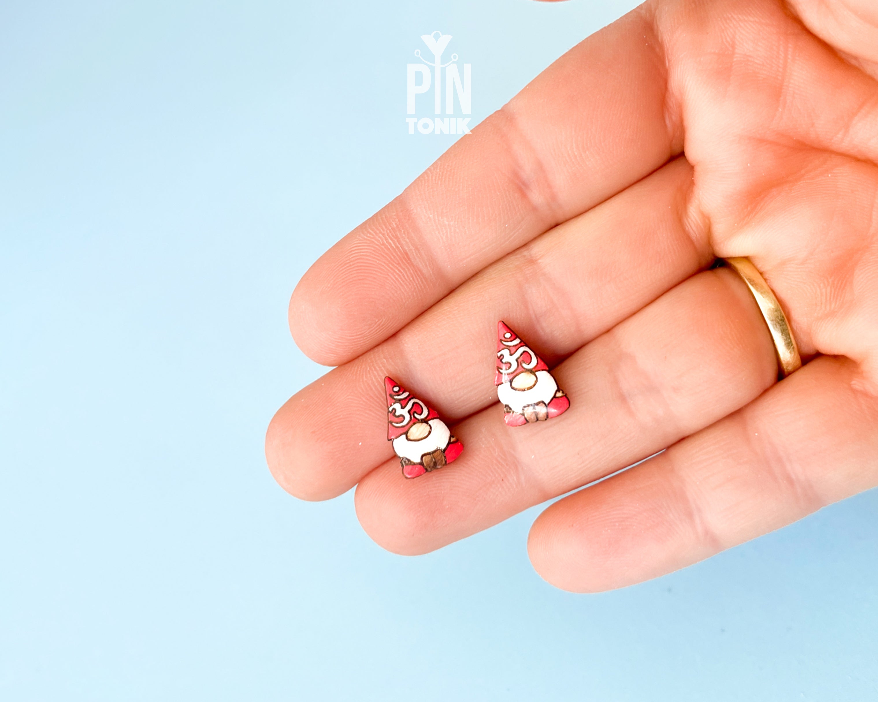 A close-up of a hand holding two small gnome-shaped stud earrings against a light blue background. 